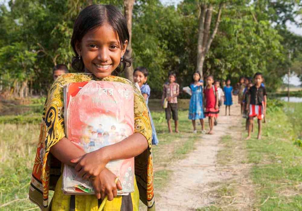 स्कूल की किताबें थामे मुस्कुराती एक लड़की, पीछे गाँव की पगडंडी पर चलते दूसरे बच्चे